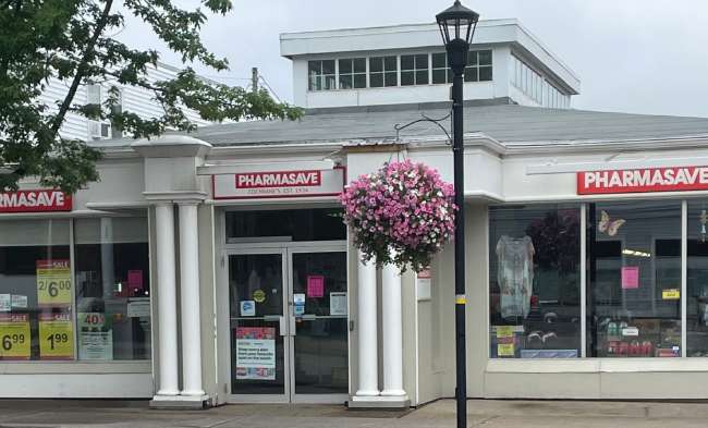 Exterior of a Pharmasave store with a hanging flower basket and sale signs in the windows.